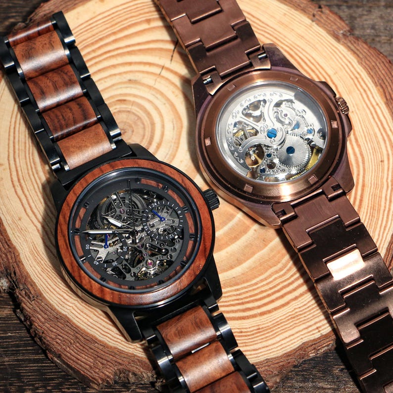 May include: Two skeleton watches with wooden and metal straps. One watch has a black case and a brown wooden bezel and strap. The other watch has a copper-coloured case and strap. The watch faces reveal the intricate inner workings.