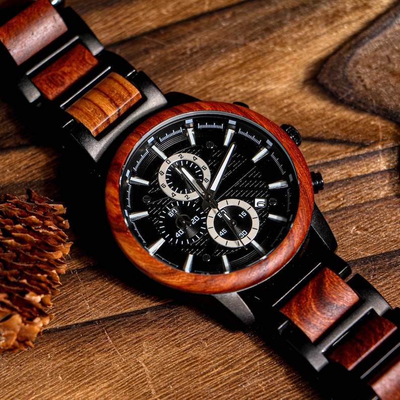 K&ouml;nnte beinhalten: Eine Holzarmbanduhr mit schwarzem Zifferblatt und brauner Holzl&uuml;nette. Die Uhr hat ein schwarzes Armband mit Holzakzenten. Das Zifferblatt der Uhr hat wei&szlig;e Zeiger und Markierungen sowie zwei kleinere Zifferbl&auml;tter. Die Uhr wird auf einer Holzoberfl&auml;che pr&auml;sentiert.