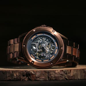 May include: A copper-coloured skeleton watch with a visible mechanical movement. The watch has a round face with a dark inner ring and a matching copper-coloured strap. The watch is displayed on a wooden surface.
