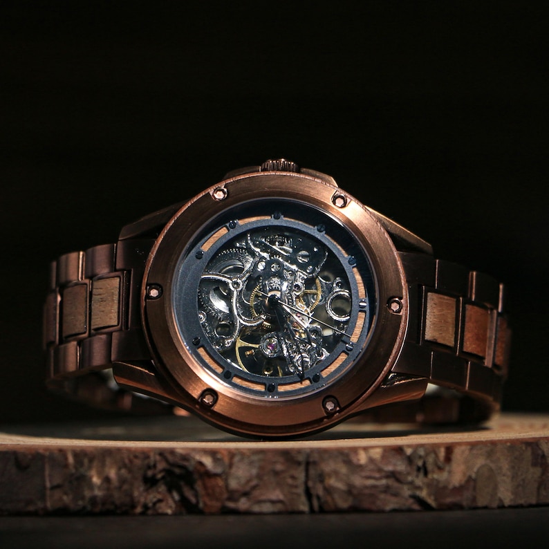May include: A copper-coloured skeleton watch with a visible mechanical movement. The watch has a round face with a dark inner ring and a matching copper-coloured strap. The watch is displayed on a wooden surface.