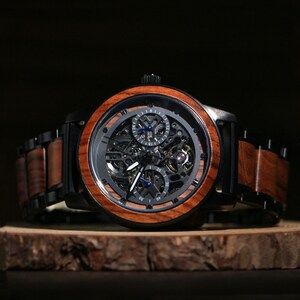 May include: A wooden wristwatch with a black face and a brown wooden bezel. The watch has a skeleton design, revealing the inner workings. The band is a combination of black metal and brown wood. The watch is displayed on a wooden surface.