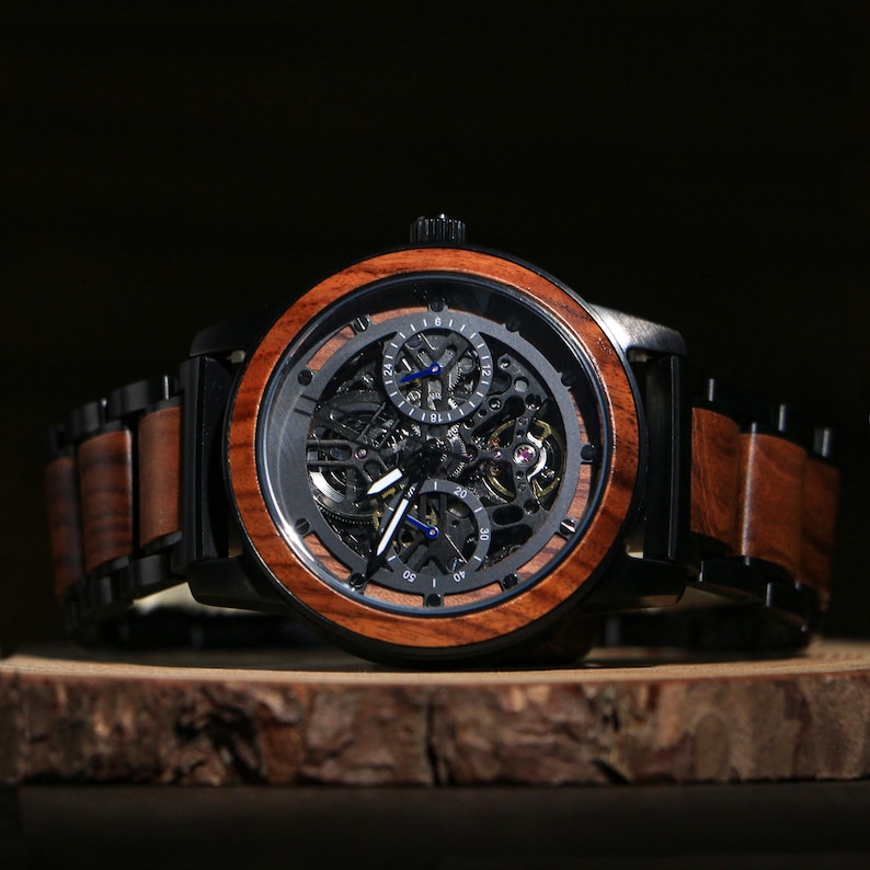 May include: A wooden wristwatch with a black face and a brown wooden bezel. The watch has a skeleton design, revealing the inner workings. The band is a combination of black metal and brown wood. The watch is displayed on a wooden surface.