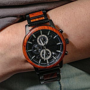 K&ouml;nnte beinhalten: Eine Holzarmbanduhr mit schwarzem Zifferblatt und brauner Holzl&uuml;nette. Die Uhr hat drei Hilfszifferbl&auml;tter und ein schwarzes Metallarmband. Die Uhr befindet sich am Handgelenk einer Person.