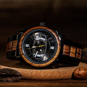 K&ouml;nnte beinhalten: Eine schwarze Armbanduhr mit Holzl&uuml;nette und -armband. Das Zifferblatt der Uhr ist schwarz mit wei&szlig;en und gelben Akzenten. Die Uhr liegt auf einer Holzoberfl&auml;che.