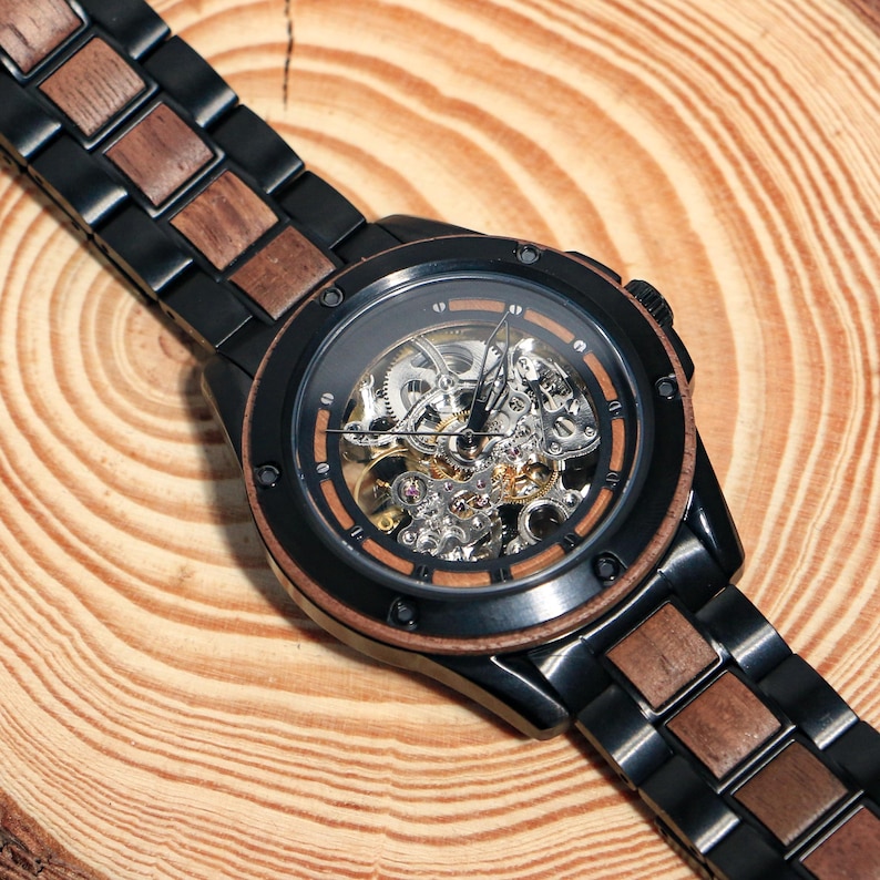 May include: A black and brown wooden wristwatch with a skeleton dial, revealing the internal mechanisms. The watch has a black case and a band with alternating wood and black links. The watch is presented on a wooden surface.