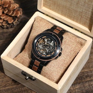 May include: A black and wood-toned skeleton watch with a visible mechanical movement, presented in a wooden box with a burlap-like cushion. The watch features a black bezel and a matching black band with wood-toned accents.