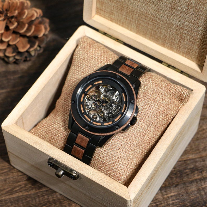 May include: A black and wood-toned skeleton watch with a visible mechanical movement, presented in a wooden box with a burlap-like cushion. The watch features a black bezel and a matching black band with wood-toned accents.