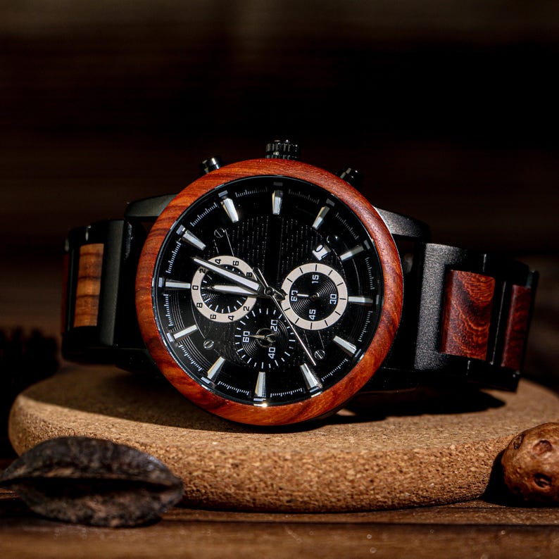 K&ouml;nnte beinhalten: Eine Holzarmbanduhr mit schwarzem Zifferblatt und brauner Holzl&uuml;nette. Die Uhr hat ein schwarzes Armband mit Holzakzenten. Die Uhr wird auf einer Korkoberfl&auml;che pr&auml;sentiert.
