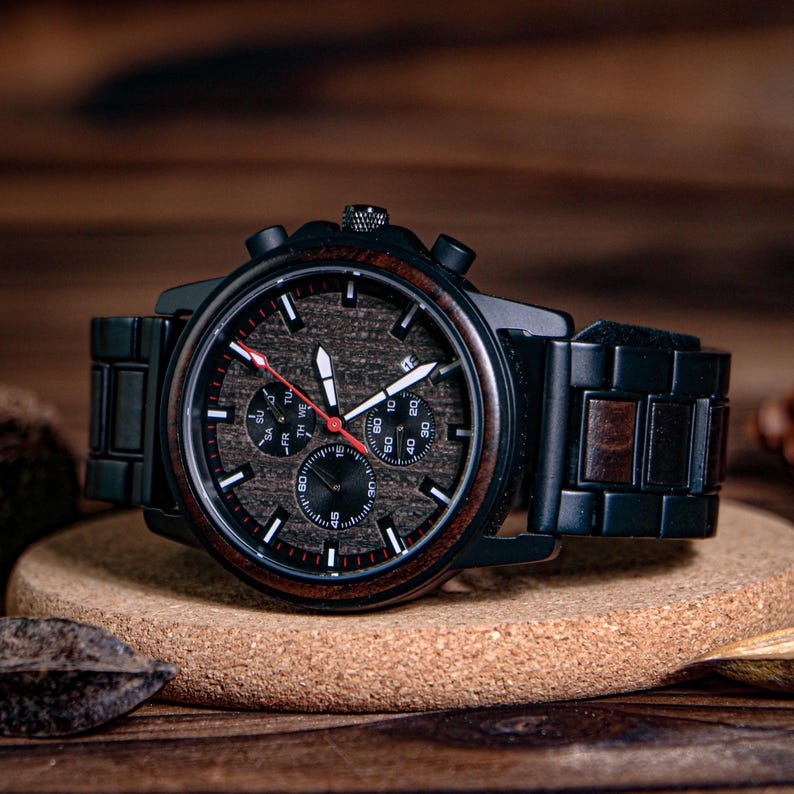 K&ouml;nnte beinhalten: Eine schwarze Holzarmbanduhr mit einem dunklen Holzmaserungszifferblatt und einem roten Sekundenzeiger. Die Uhr hat ein schwarzes Metallgeh&auml;use und -armband mit Holzmaserungsakzenten. Die Uhr wird auf einer Korkoberfl&auml;che pr&auml;sentiert.