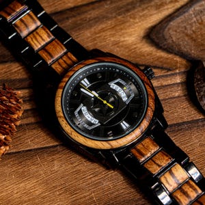 K&ouml;nnte beinhalten: Eine Holzarmbanduhr mit schwarzem Zifferblatt und Holzl&uuml;nette. Die Uhr hat ein schwarzes Metallarmband mit Holzakzenten. Die Zeiger der Uhr sind gelb. Die Uhr liegt auf einer Holzoberfl&auml;che.