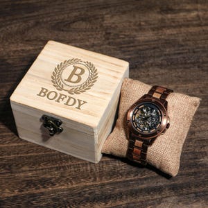 May include: A wooden watch box, engraved with "BOFDY" and a "B" initial within a laurel wreath, sits beside a bronze skeleton watch on a burlap cushion. The watch features a visible mechanical movement and a bronze strap.