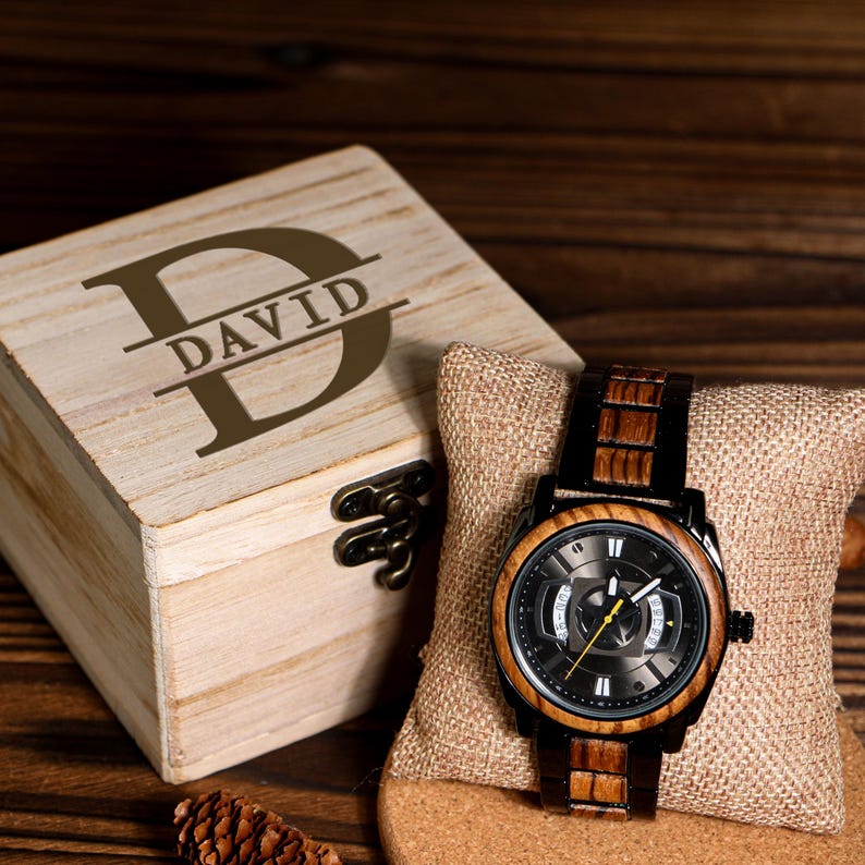 K&ouml;nnte beinhalten: Eine Holzuhr mit schwarzem Zifferblatt und Holzarmband liegt auf einem Leinenkissen. Die Uhr wird neben einer hellen Holzkiste mit dem eingravierten Namen "DAVID" auf dem Deckel pr&auml;sentiert. Die Uhr hat eine schwarze L&uuml;nette und einen gelben Sekundenzeiger.