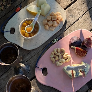 May include: Two ceramic serving boards with food and drinks on a wooden table. One board is white with melon, cheese cubes, and a bowl of jam. The other is pink with figs, cashews, and blue cheese. Two silver goblets are also present.