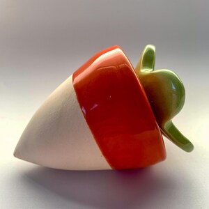 May include: A ceramic strawberry-shaped object featuring a matte white pointed base, a glossy orange midsection, and a smooth, shiny green leaf-shaped top. The piece has a conical form with a rounded, organic shape and a reflective surface finish.