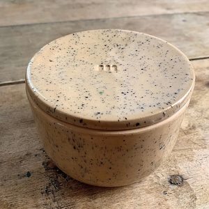 May include: A speckled beige ceramic container with a lid. The lid has three small, evenly spaced indentations and is covered in small black speckles. The container's body also features the same speckled pattern. The container is round and sits on a wooden surface.