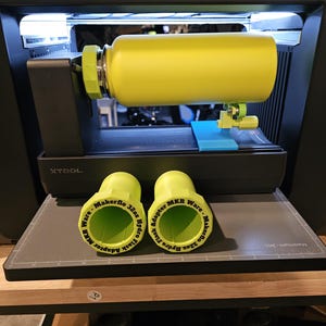 May include: A bright yellow water bottle mounted on an xTool rotary attachment inside a laser machine. Two 3D-printed lime green adapters are placed in the foreground, each labeled with the text Makerflo 32oz Hydro Flask Adapter MKR Ware.