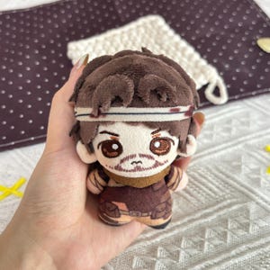 Puede incluir: Una pequeña muñeca de peluche que representa a un personaje con cabello castaño, una cinta blanca y rasgos faciales bordados, incluyendo ojos marrones y barba. La muñeca viste un conjunto marrón con cuello color canela y bolsillos utilitarios.