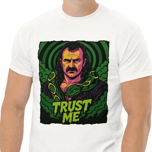 Può includere: T-shirt bianca con una grafica colorata di un uomo con i baffi e un serpente, circondato da vortici verdi e neri. Il testo "TRUST ME" è sotto l'immagine. La maglietta è indossata da una persona.