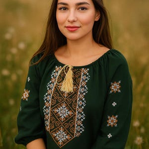May include: A dark green blouse with detailed embroidery in white and orange, featuring a round neckline and three-quarter sleeves. The blouse has a tassel detail and is set against a blurred background of green and yellow, suggesting a field.