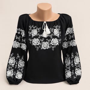 Könnte beinhalten: Schwarze Bluse mit weißer Blumenstickerei. Das langärmlige Oberteil hat einen Rundhalsausschnitt und ein weißes Quastendetail. Das florale Design konzentriert sich auf den Ausschnitt und die Ärmel.