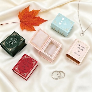 Könnte beinhalten: Eine Sammlung miniaturer ringförmiger Schatullen in Buchform in verschiedenen Farben, darunter Grün, Rot und Rosa, mit goldenen Akzenten. Einige Schatullen sind geöffnet und zeigen Ringfächer. Die Schatullen sind mit Namen und Daten personalisiert. Eine silberne Halskette mit Herzanhänger und zwei silberne Ringe sind ebenfalls vorhanden.