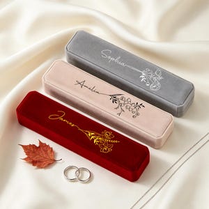 May include: Three rectangular velvet jewellery boxes in grey, light pink, and deep red. Each box features a custom name in cursive script and a floral line art illustration. The grey box says Sophia, the pink box says Amelia, and the red box says James.