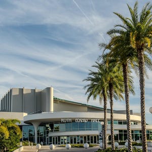 Puede incluir: Vista exterior del Ruth Eckerd Hall, un edificio moderno con fachada curva y grandes ventanales. Palmeras bordean el frente, y una bandera estadounidense ondea bajo un cielo parcialmente nublado.