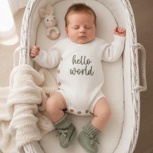 Pode incluir: Um bebé a dormir veste um body de malha branco de manga comprida com o texto hello world bordado a cinzento, combinado com botinhas de malha verde sálvia. Um pequeno chocalho de coelho em croché creme repousa num berço de vime branco.