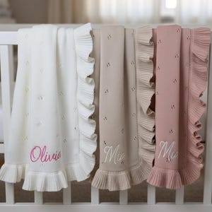 Può includere: Tre coperte per bambini in crema, beige e rosa, ciascuna con un bordo a balze e un nome ricamato. La coperta color crema ha il nome "Olivia" in rosa. Le coperte beige e rosa hanno il nome "Mia" in bianco. Le coperte sono esposte su una culla bianca.