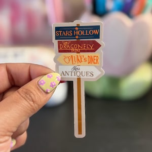 May include: A sticker featuring a signpost with arrows pointing to Stars Hollow, Dragonfly Inn, Luke's Diner, and Kim's Antiques. The signpost has a tan pole and white, red, yellow, and blue arrows. The text is in a serif font.