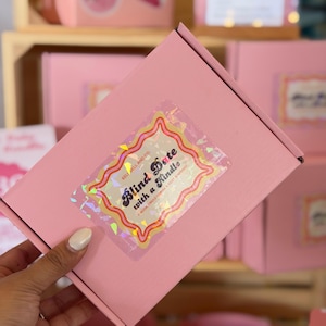 May include: A pink box with a holographic sticker that reads "Blind Date with a Kindle". The sticker has a decorative border with red and yellow accents. The box is held by a hand, with other boxes visible in the background.