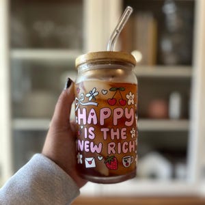 May include: A glass jar with a bamboo lid and a clear straw, filled with a beverage. The jar is decorated with the phrase "HAPPY IS THE NEW RICH" in pink lettering, along with floral and fruit illustrations.