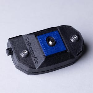 May include: A black, rectangular 3D-printed object with a blue square insert and silver screws. The text "Takomoto Co" is printed on the side. The object has a small, black, rectangular protrusion on one end.