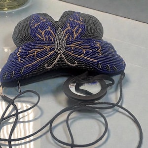 May include: A beaded butterfly-shaped purse in black and blue with gold accents. The purse has a black strap and a small, round mirror attached. The butterfly design is detailed with small beads, creating a textured surface. The purse is likely a vintage or handmade item.