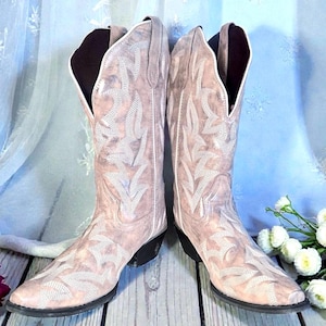 May include: A pair of light pink cowboy boots with intricate white stitching. The boots have a pointed toe, a black heel, and a tall shaft. The design features a floral pattern. The boots are displayed against a light blue backdrop.