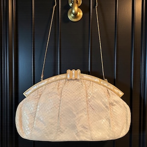 May include: A vintage, light beige clutch with a snakeskin pattern. The purse has a gold-toned frame and clasp, and a delicate gold chain strap. The bag is hanging against a black background.