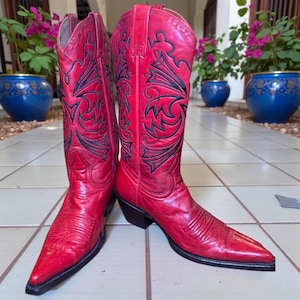 May include: A pair of red cowboy boots with black embroidered floral designs. The boots have pointed toes and black soles. The leather boots are displayed on a tiled surface, with potted plants in the background.