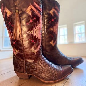 May include: A pair of dark brown leather cowboy boots featuring a textured snakeskin-patterned lower section, a pointed toe, and a stacked heel. The upper shaft is decorated with intricate geometric embroidery in pink and tan, plus a brown tassel.