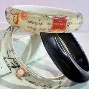May include: Three bangles are stacked, showcasing a variety of designs. Two are translucent with a cream-colored background, featuring script and cameo details. The third is solid black. The bangles are likely for the wrist.