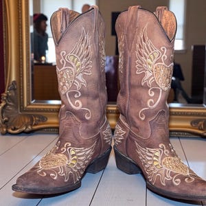 May include: A pair of brown cowboy boots with intricate embroidered designs. The boots feature wing and floral patterns in gold and cream thread. The leather boots have pointed toes and angled heels, suitable for Western wear.