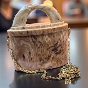 May include: A rectangular handbag with a curved handle and a gold chain strap. The bag is made of a light brown material with dark brown swirling patterns. The handle and top trim are a lighter shade of brown.