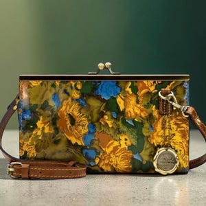 May include: A rectangular floral print crossbody bag with a brown leather strap and a clasp closure. The bag features a colorful pattern of yellow, blue, and green flowers. A small, round, gold-colored emblem hangs from the strap.