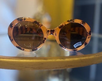 Könnte beinhalten: Ovale Sonnenbrille mit Schildpattmuster. Die Fassungen sind hellorange mit dunkelbraunen Flecken. Die Gläser sind dunkelbraun. Die Sonnenbrille hat einen UV400-Schutzaufkleber.