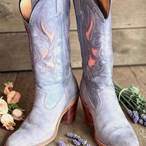 May include: A pair of blue-grey cowboy boots with decorative red stitching. The boots have a pointed toe, wooden heels, and are displayed on a wooden surface with lavender and flowers.