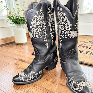 May include: A pair of black leather cowboy boots with intricate cream-colored floral embroidery. The boots have pointed toes and stacked heels. The decorative stitching extends up the shafts, adding a touch of Western flair to the footwear.