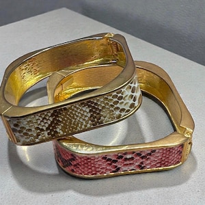 May include: Two gold-toned square bangles with snake print detailing. One bangle features a brown, white, and tan pattern, while the other displays a pink, black, and white pattern. The bracelets are stacked on top of each other.