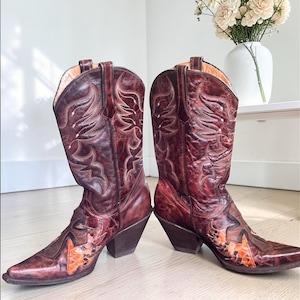 May include: A pair of burgundy cowboy boots with pointed toes and angled heels. The boots feature intricate stitching and decorative patterns. The toe and heel areas have a contrasting orange and brown design. The boots are made of leather.