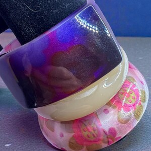 May include: Three stacked bangles. The top bangle is a deep purple with a glossy finish. The middle bangle is a light beige. The bottom bangle is pink with a floral pattern in pink and gold.