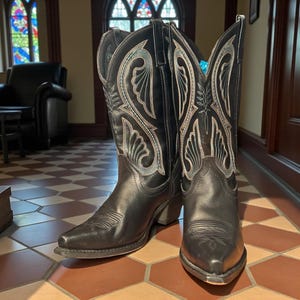 May include: A pair of black leather cowboy boots with intricate white and teal stitching. The boots have pointed toes, stacked heels, and decorative patterns on the shafts. They are standing on a tiled floor.