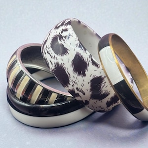 May include: Four stacked bangles in a variety of designs. One bangle has a white base with brown spots, another has black and white stripes, and a third has a black and white design with a gold band. The fourth bangle is black and white.